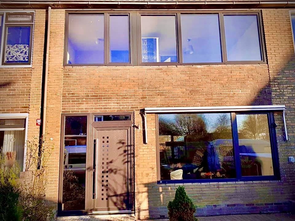 Brązowe drzwi wejściowe z szybą i symetrycznym wzorem kwadratów, zamontowane obok dużego okna przesuwnego na tle ceglanej ściany budynku.