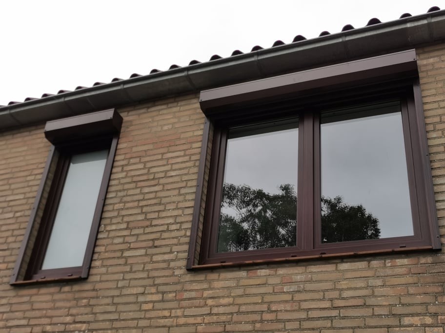 Brązowe okna PCV z roletami zewnętrznymi w kolorze dopasowanym do ramy, zamontowane w ścianie z cegły klinkierowej.