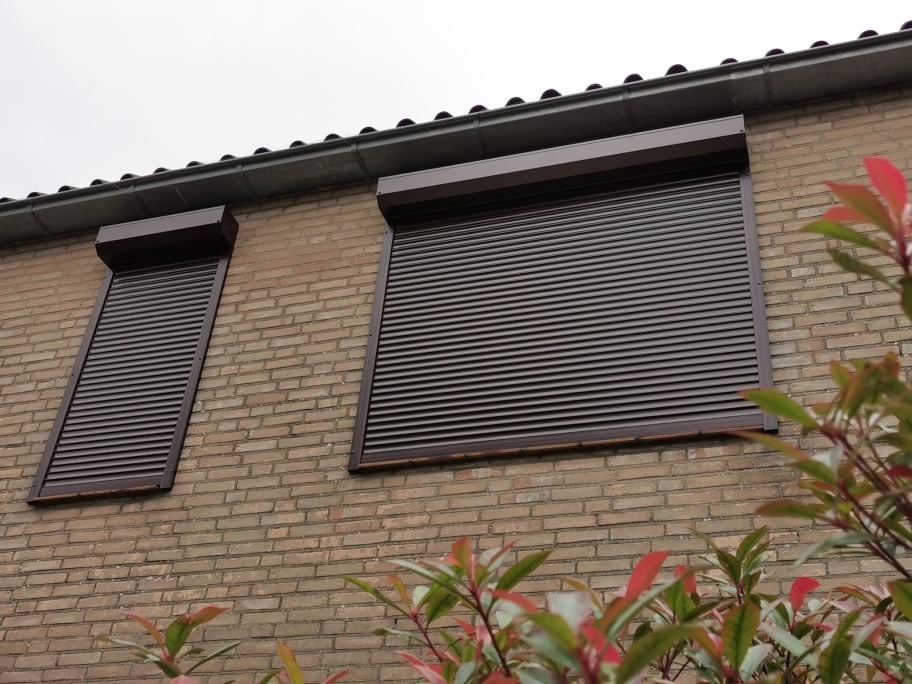 Dwa okna na ceglanej ścianie budynku, zabezpieczone brązowymi roletami zewnętrznymi, częściowo zasłonięte przez krzew z czerwonymi liśćmi.