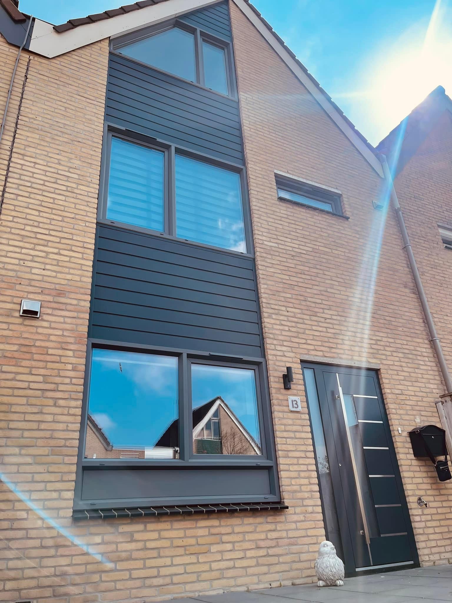Elewacja domu z cegły klinkierowej z szarymi oknami z refleksami nieba i nowoczesnymi drzwiami wejściowymi z numerem 13, dekoracyjna figurka ptaka przed drzwiami.
