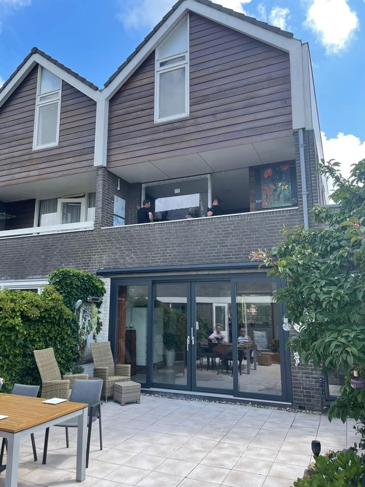 Montaż okna na balkonie w dwupiętrowym domu z drewnianą elewacją, widoczne dwie osoby przy pracy, na dole taras z szarymi przesuwnymi drzwiami i meblami ogrodowymi.