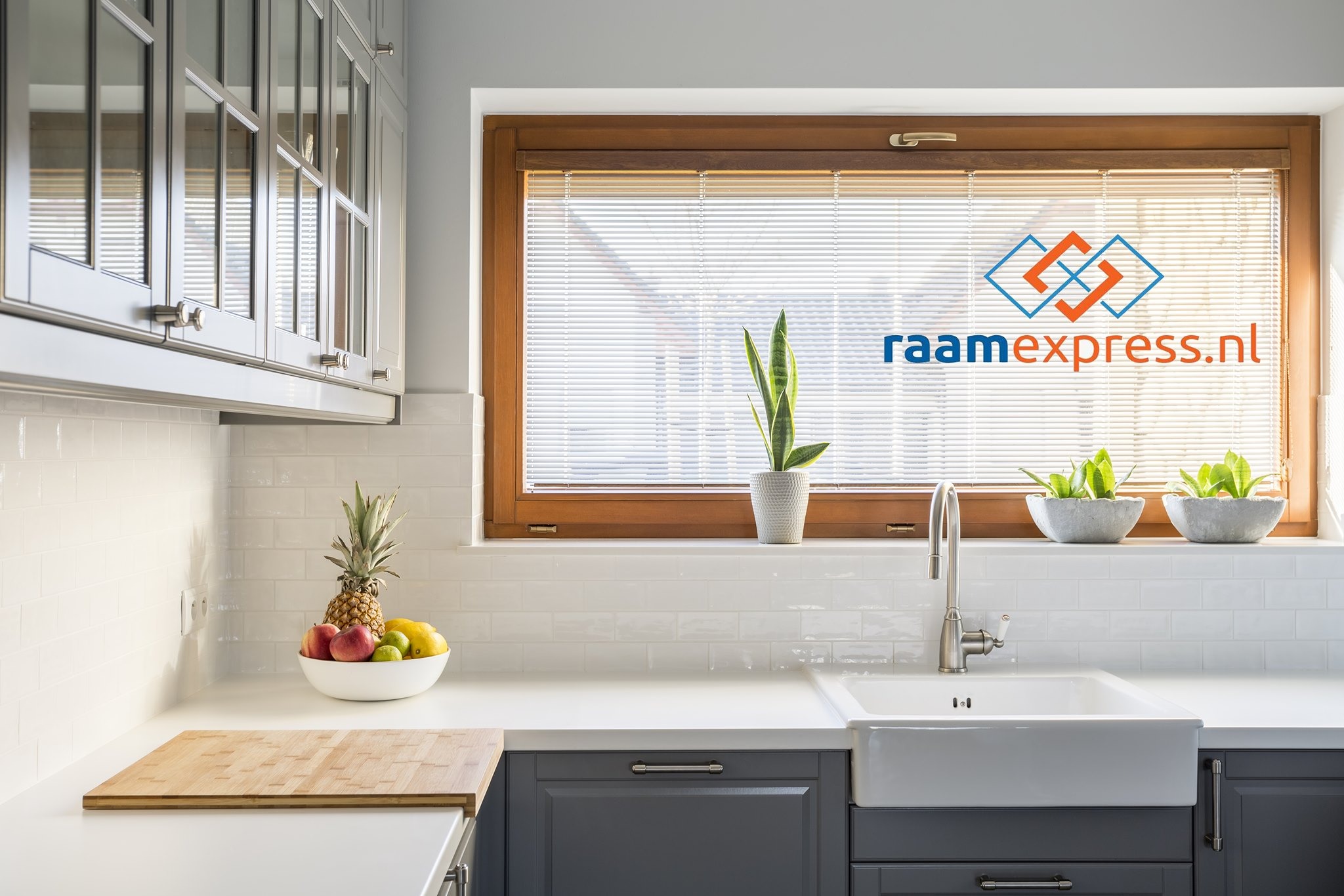 Kuchnia z oknem w drewnianej ramie, zasłoniętym poziomymi żaluzjami, na tle których widnieje logo firmy raamexpress.nl. Na parapecie doniczki z roślinami, poniżej zlew ceramiczny i blat z owocami.