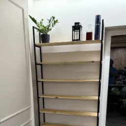 Ręcznie wykonany regał metalowy z drewnianymi półkami, umieszczony we wnętrzu obok białych drzwi, z dekoracjami na górnej półce.