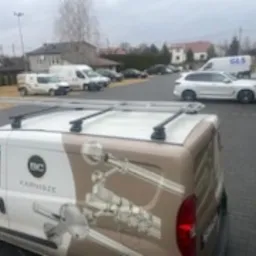 Samochód dostawczy z grafiką przedstawiającą elementy metalowe, prawdopodobnie balustrady lub poręcze, zaparkowany na parkingu firmy z widocznymi innymi pojazdami i budynkami w tle.