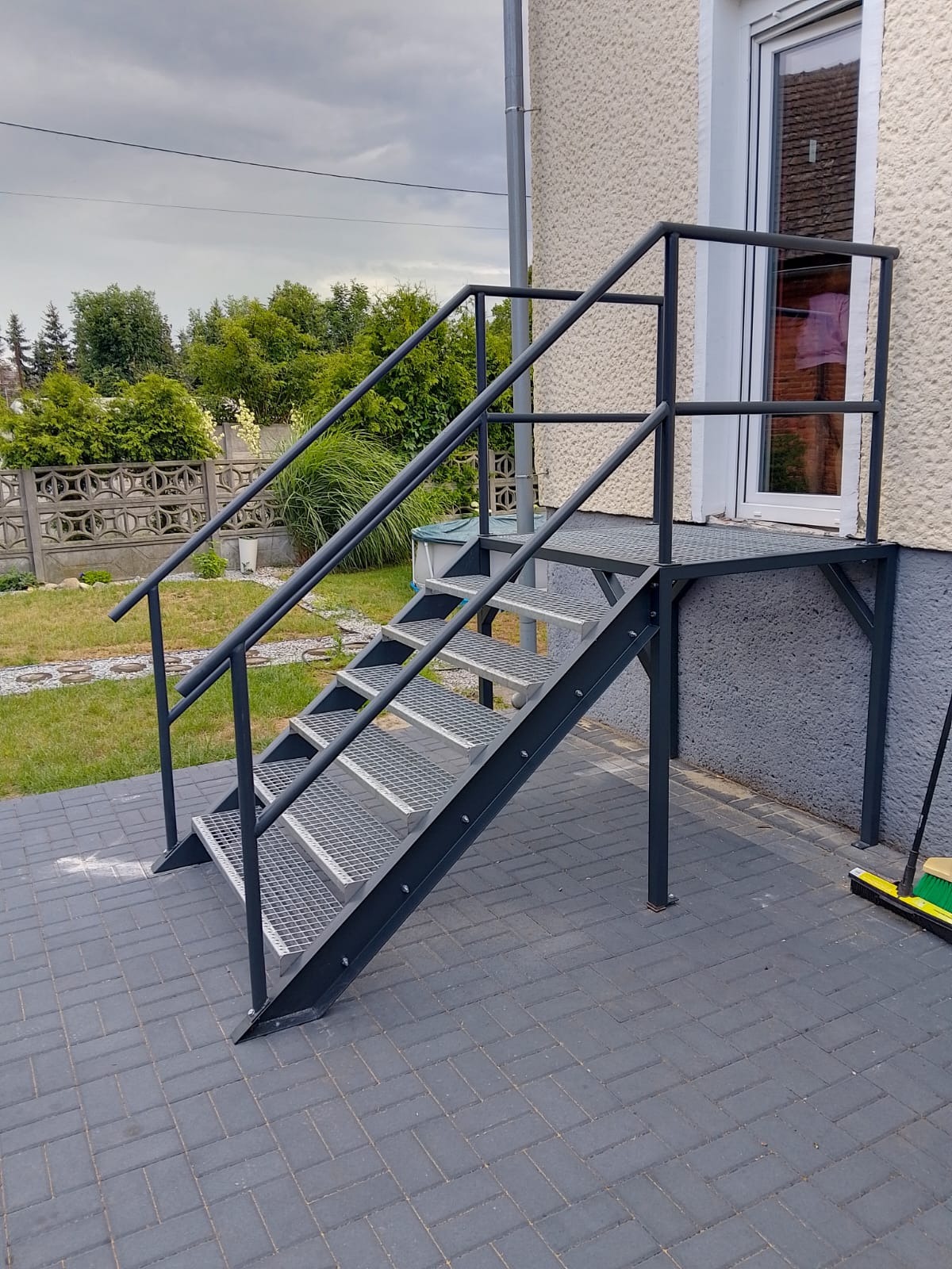 Zewnętrzne schody metalowe z poręczą, prowadzące do drzwi. Konstrukcja pomalowana na szaro, kratowe stopnie. Widoczne otoczenie domu, kostka brukowa i fragment ogrodu.