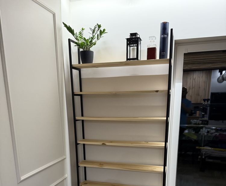 Ręcznie wykonany regał metalowy z drewnianymi półkami, umieszczony we wnętrzu obok białych drzwi, z dekoracjami na górnej półce.