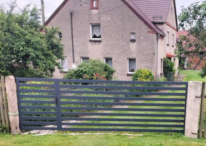 Nowoczesne, poziome ogrodzenie metalowe w kolorze grafitowym, z bramką, na tle domu z szarą elewacją i zielenią. Słupki ogrodzeniowe betonowe, lekko zniszczone.