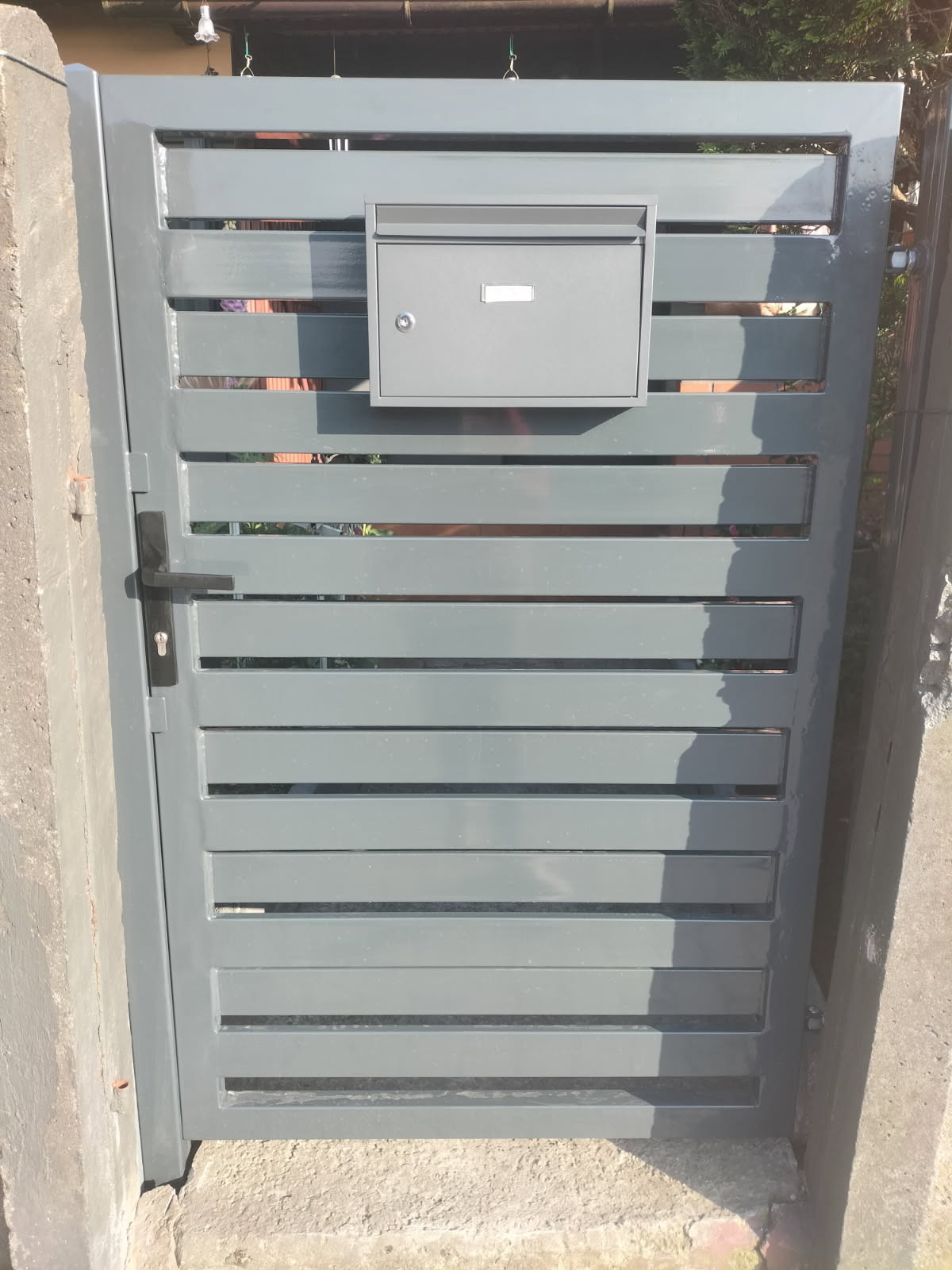 Szara, metalowa furtka z poziomymi szczeblami i skrzynką na listy. Nowoczesny design, widoczne mocowania do słupków.