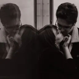 Czarno-białe zdjęcie całującej się pary, odbicie w szklanej powierzchni tworzy efekt podwojenia postaci.