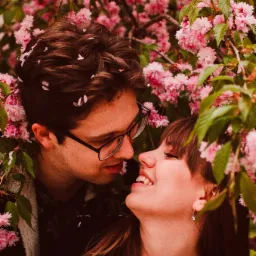 Zakochana para, on w okularach, patrzy na uśmiechniętą kobietę, otoczeni kwitnącym drzewem wiśni o różowych kwiatach.