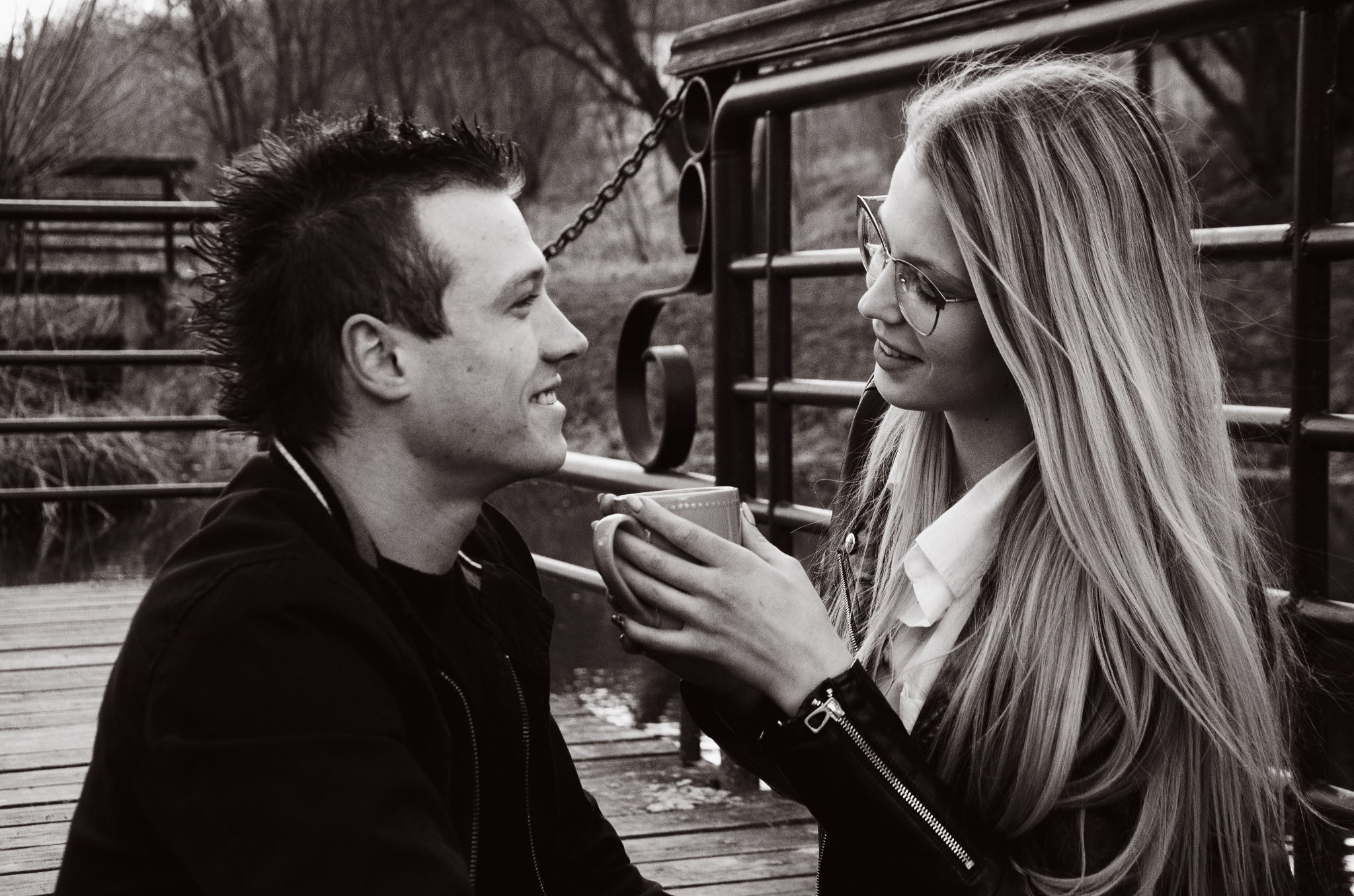Czarno-białe zdjęcie pary siedzącej na drewnianym pomoście, trzymającej razem kubek i spoglądającej na siebie z uśmiechem, z metalową barierką w tle.