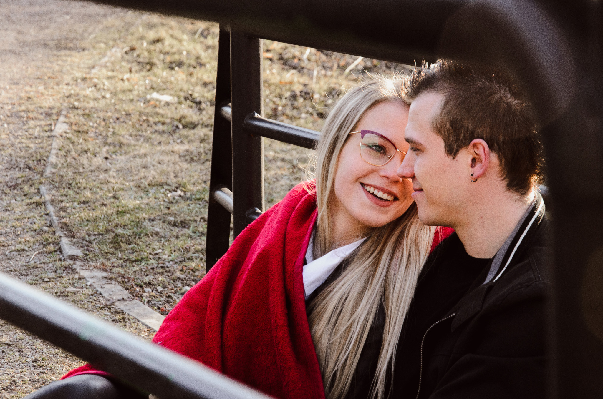 Uśmiechnięta blondynka w okularach z czerwoną chustą, obejmująca mężczyznę z kolczykiem w uchu, na tle parku widzianego przez metalową barierkę.