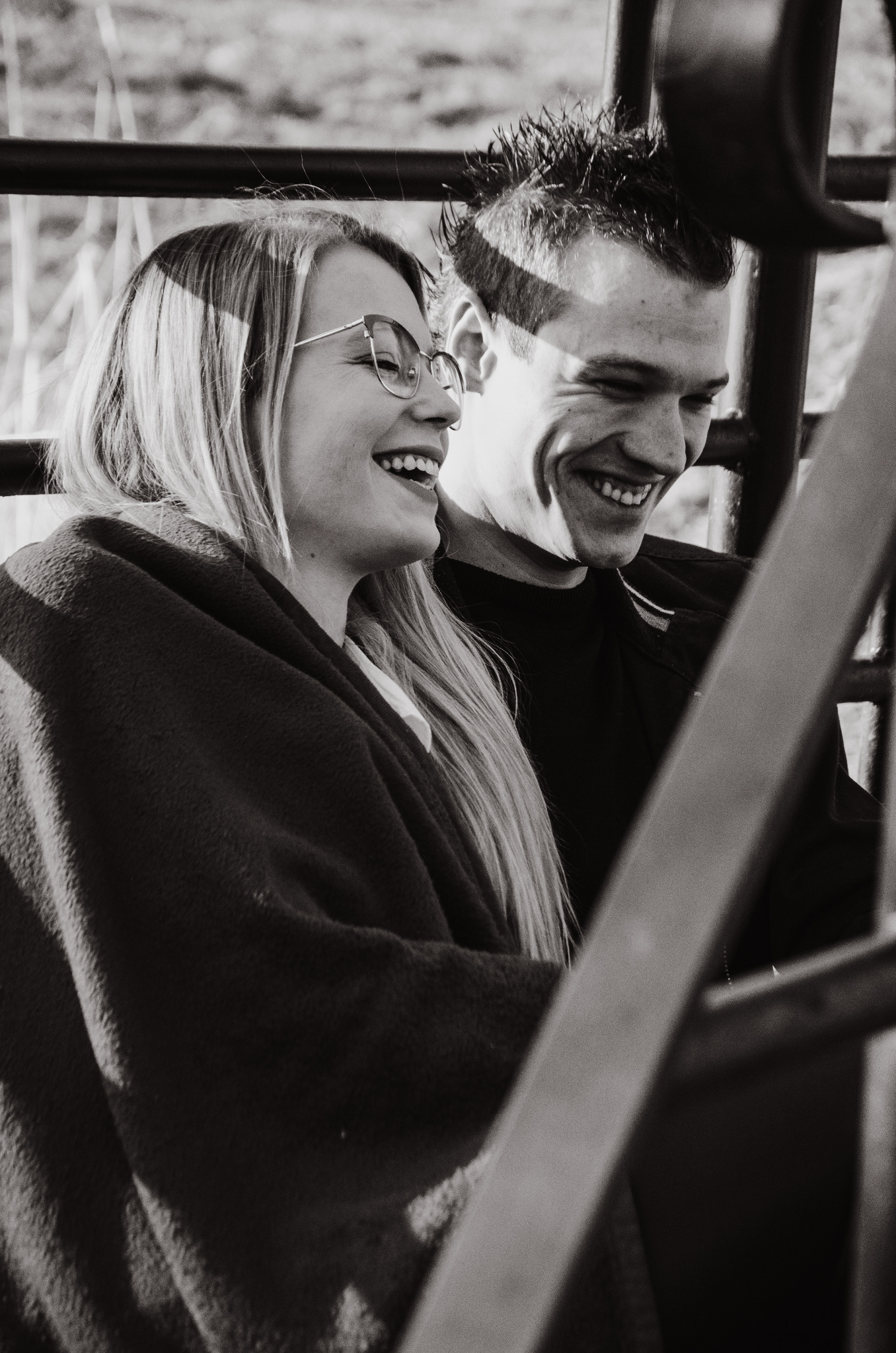 Czarno-białe zdjęcie uśmiechniętej pary, kobieta w okularach i kocu, mężczyzna z bliska, uchwycone przez metalową konstrukcję.