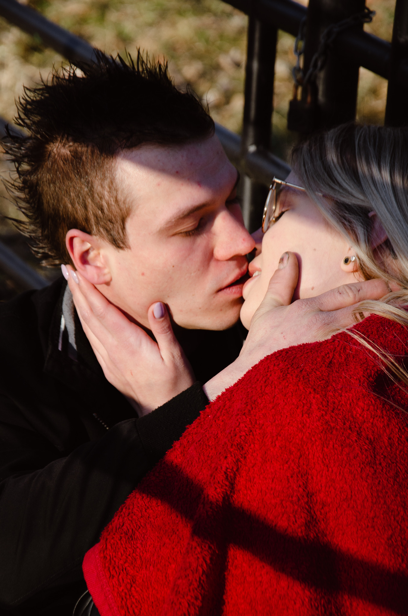 Portret całującej się pary na tle metalowego ogrodzenia z kłódkami, kobieta w okularach i czerwonym płaszczu.