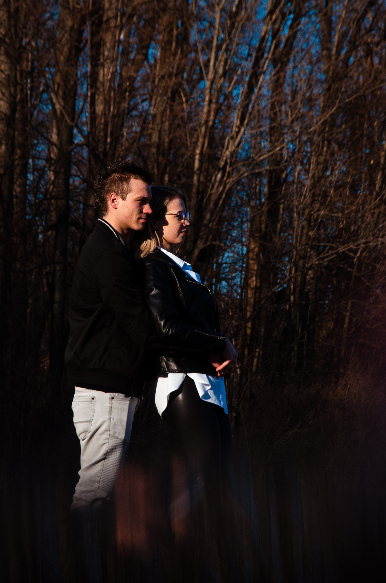Para w jesiennej scenerii: mężczyzna obejmuje kobietę w okularach, oboje patrzą w dal, drzewa bez liści w tle.
