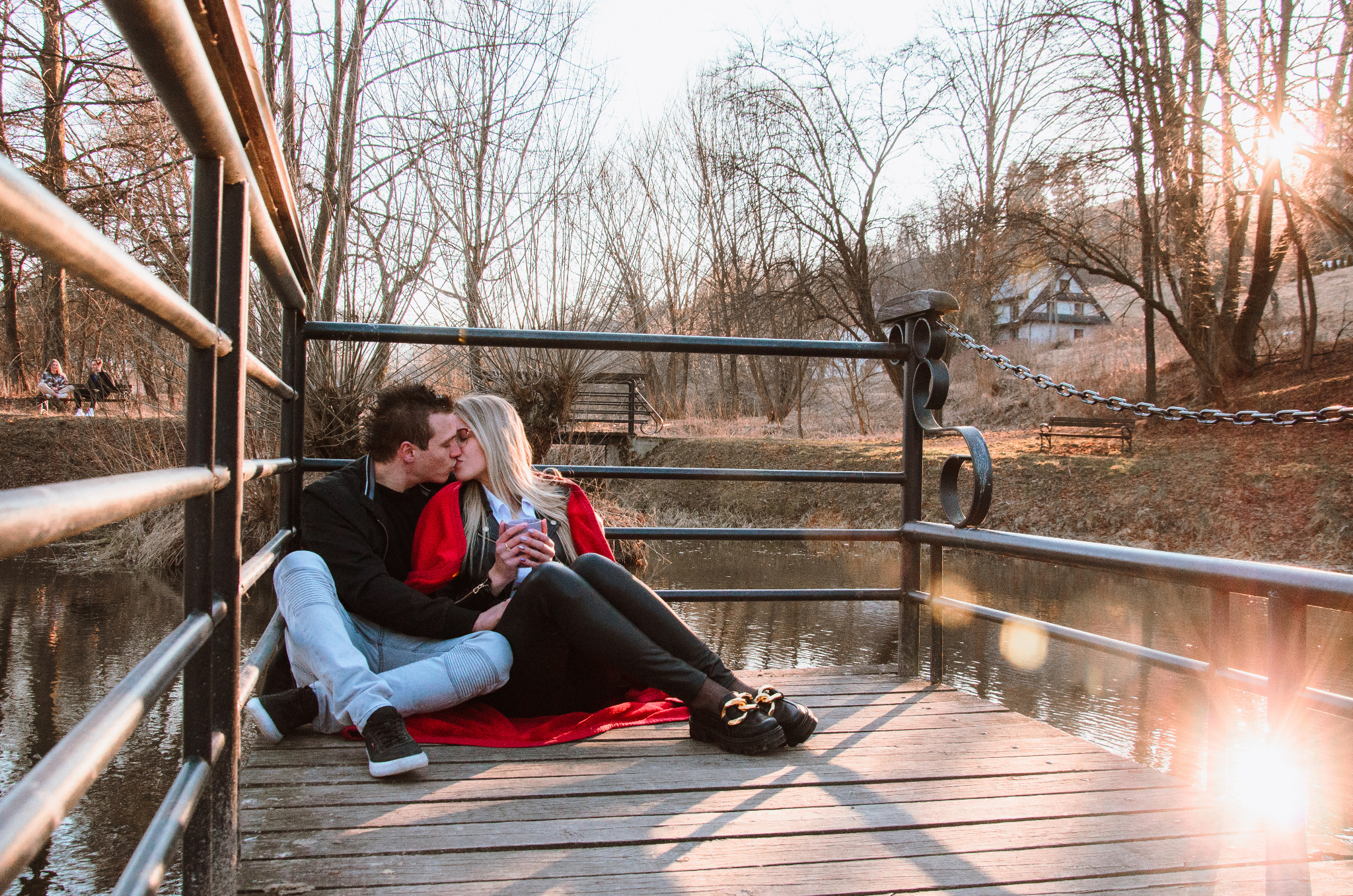 Zakochana para całuje się siedząc na drewnianym pomoście nad rzeką, okryta czerwoną narzutą, w tle widok na park z ławkami i drzewami, promienie zachodzącego słońca.
