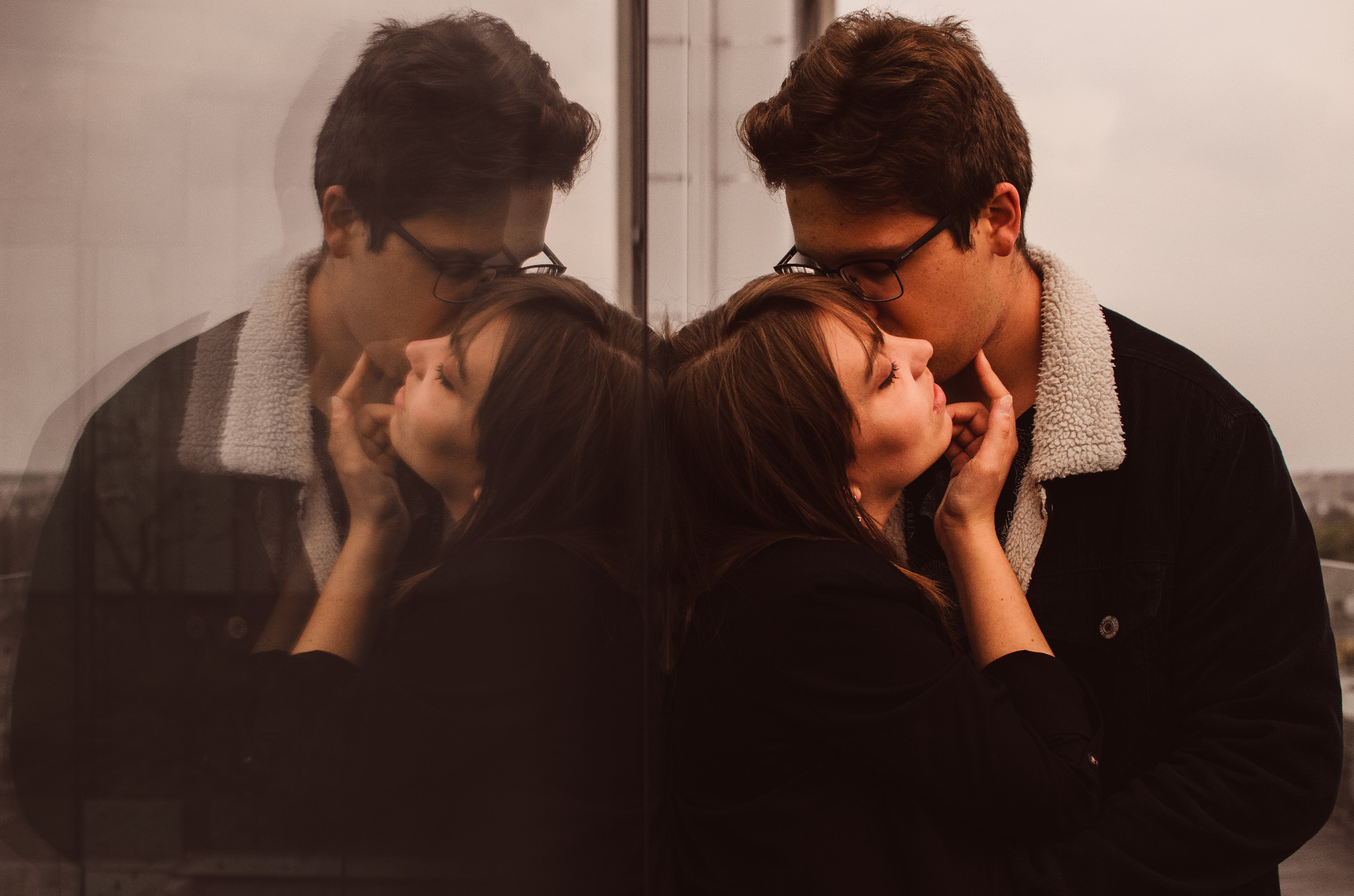 Zakochana para w kurtkach z barankiem całuje się na tle szklanej ściany, odbicie sylwetek.