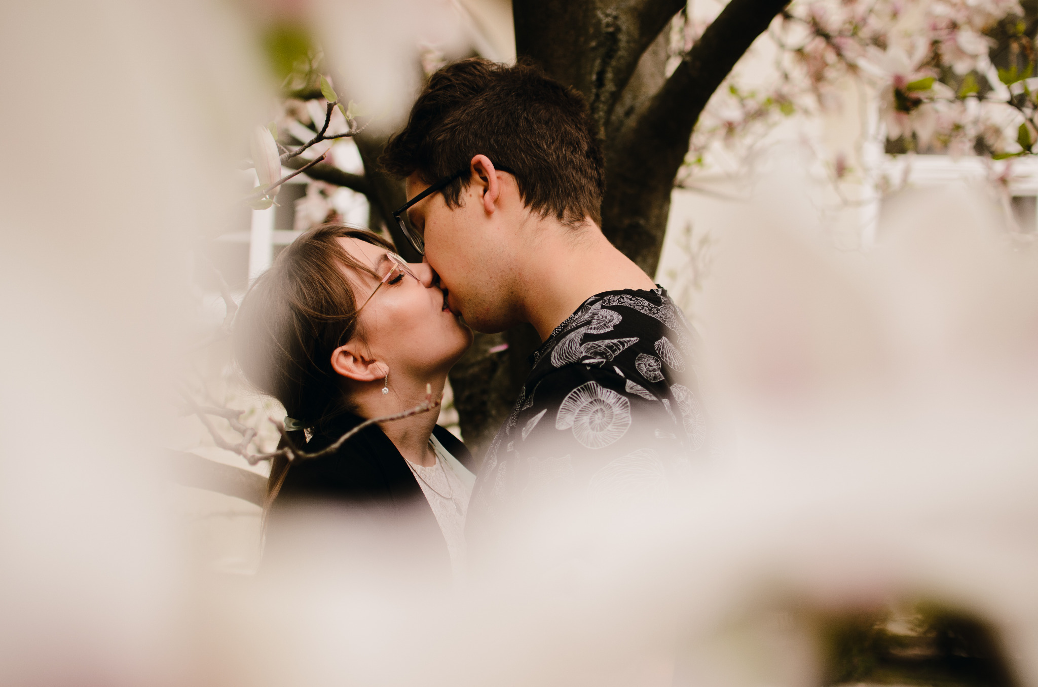 Zakochana para całuje się na tle kwitnącej magnolii, delikatne rozmycie kwiatów w pierwszym planie dodaje intymności ujęciu.