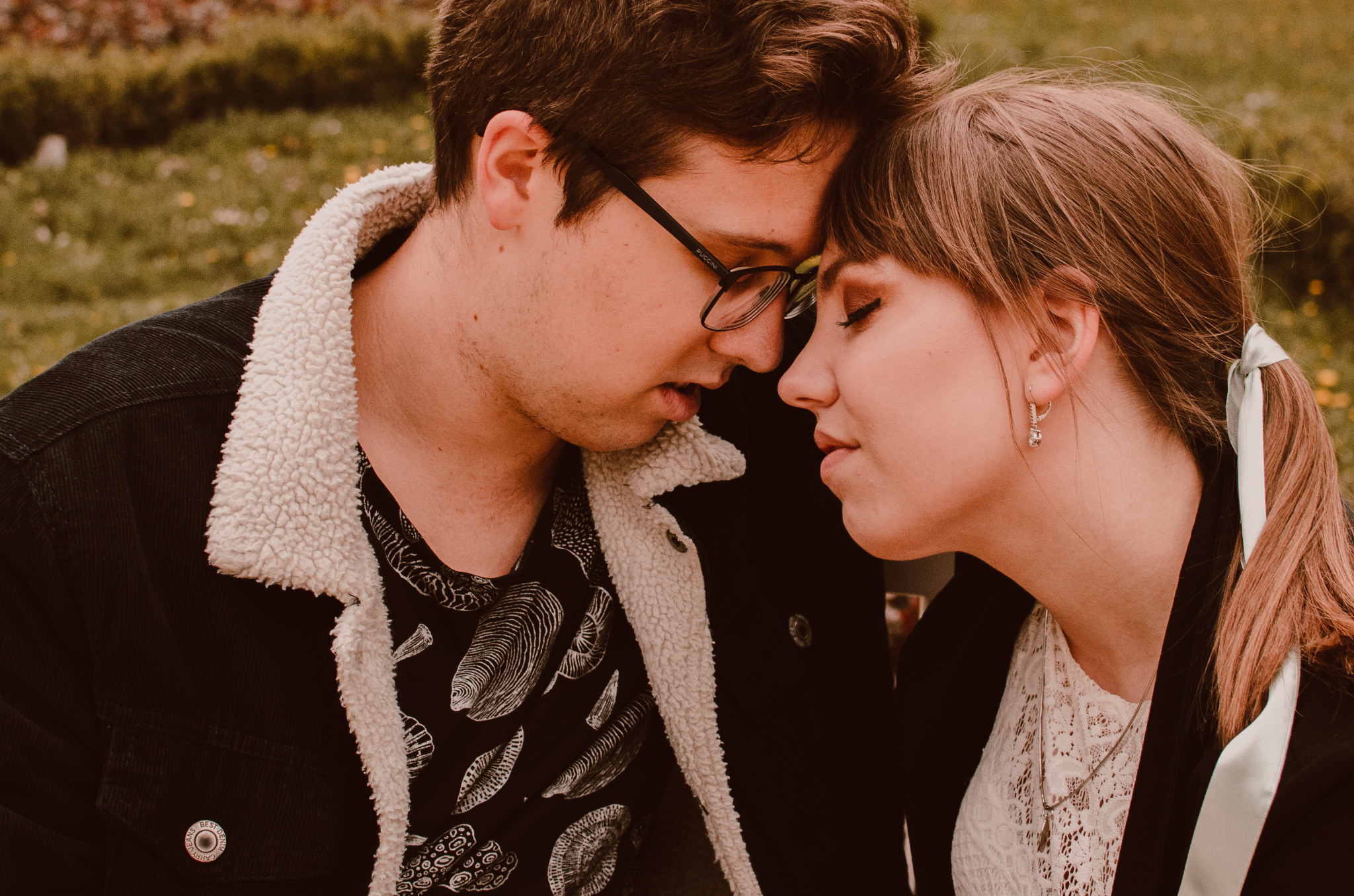 Zbliżenie na parę dotykającą się czołami; mężczyzna w okularach i czarnej kurtce z barankiem, kobieta z włosami związanymi wstążką i delikatnym makijażem oczu.