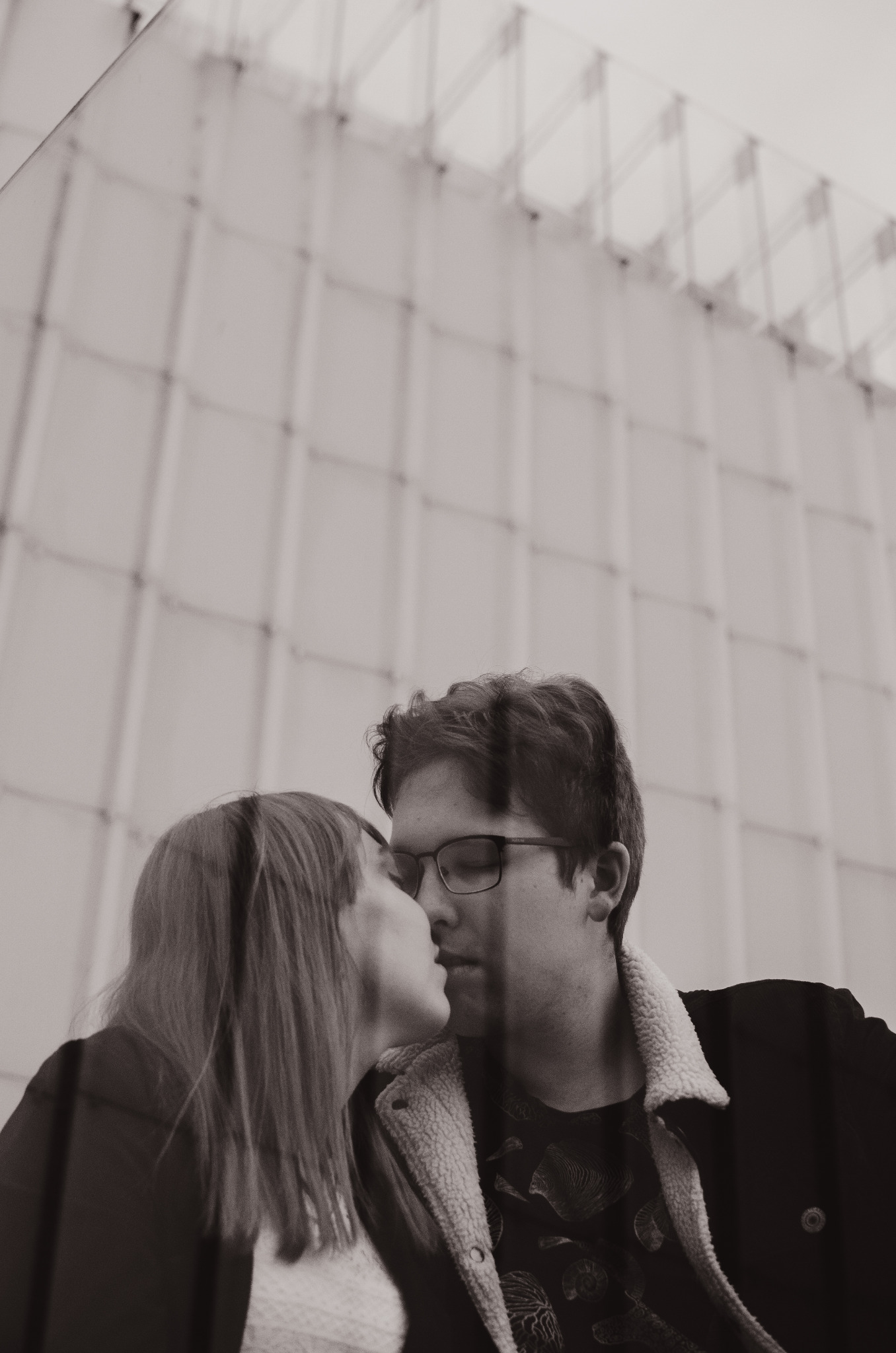 Czarno-białe zdjęcie pary całującej się na tle nowoczesnej fasady budynku z geometrycznym wzorem.