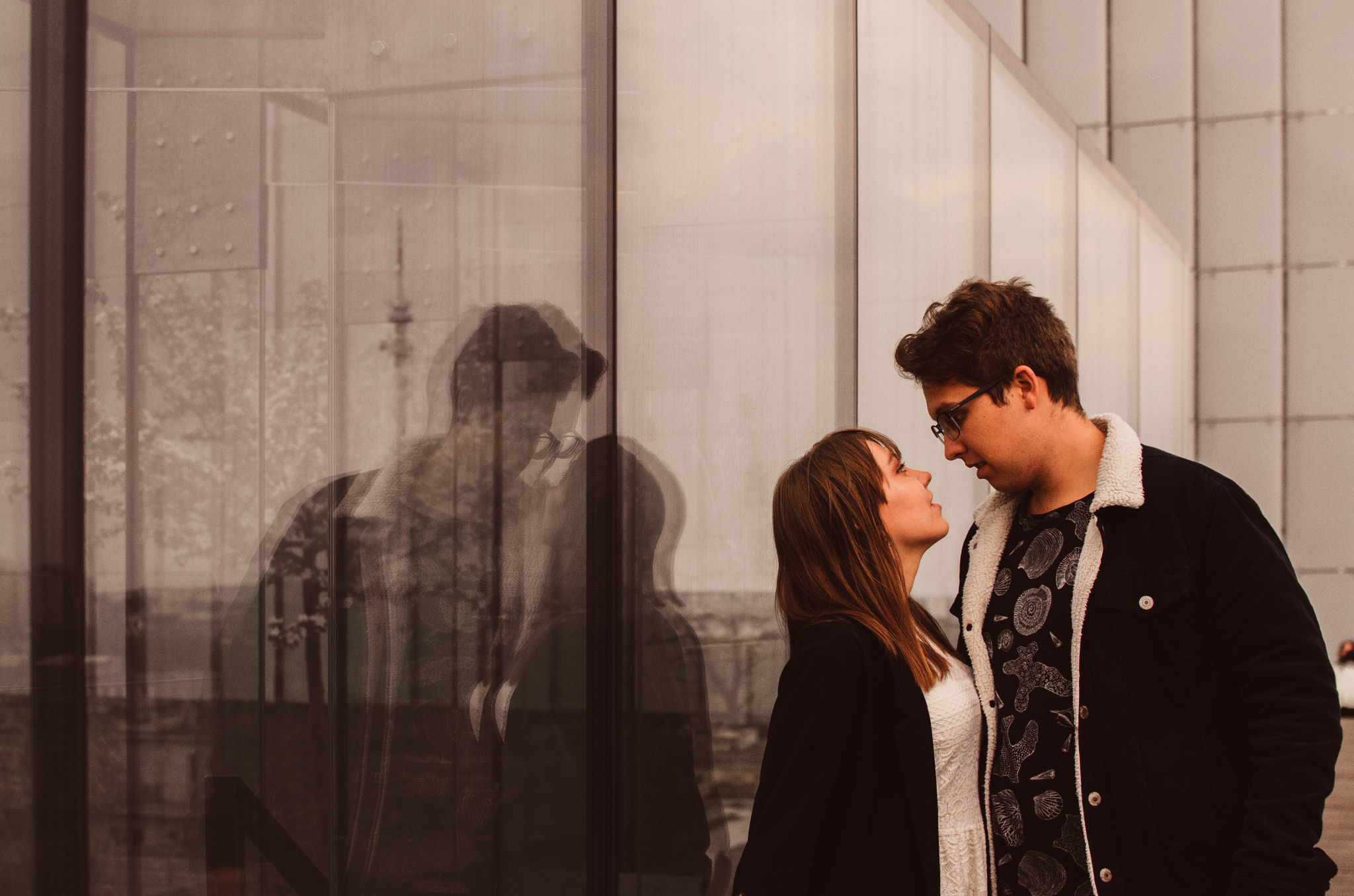 Młoda para patrzy na siebie czule na tle nowoczesnego budynku z odbijającym się w szkle krajobrazem miejskim.