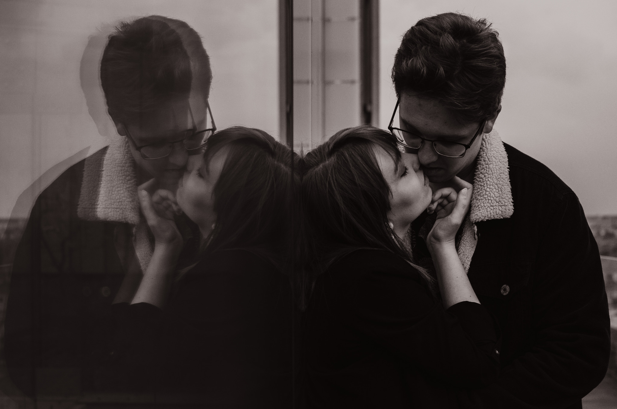 Czarno-białe zdjęcie całującej się pary, odbicie w szklanej powierzchni tworzy efekt podwojenia postaci.