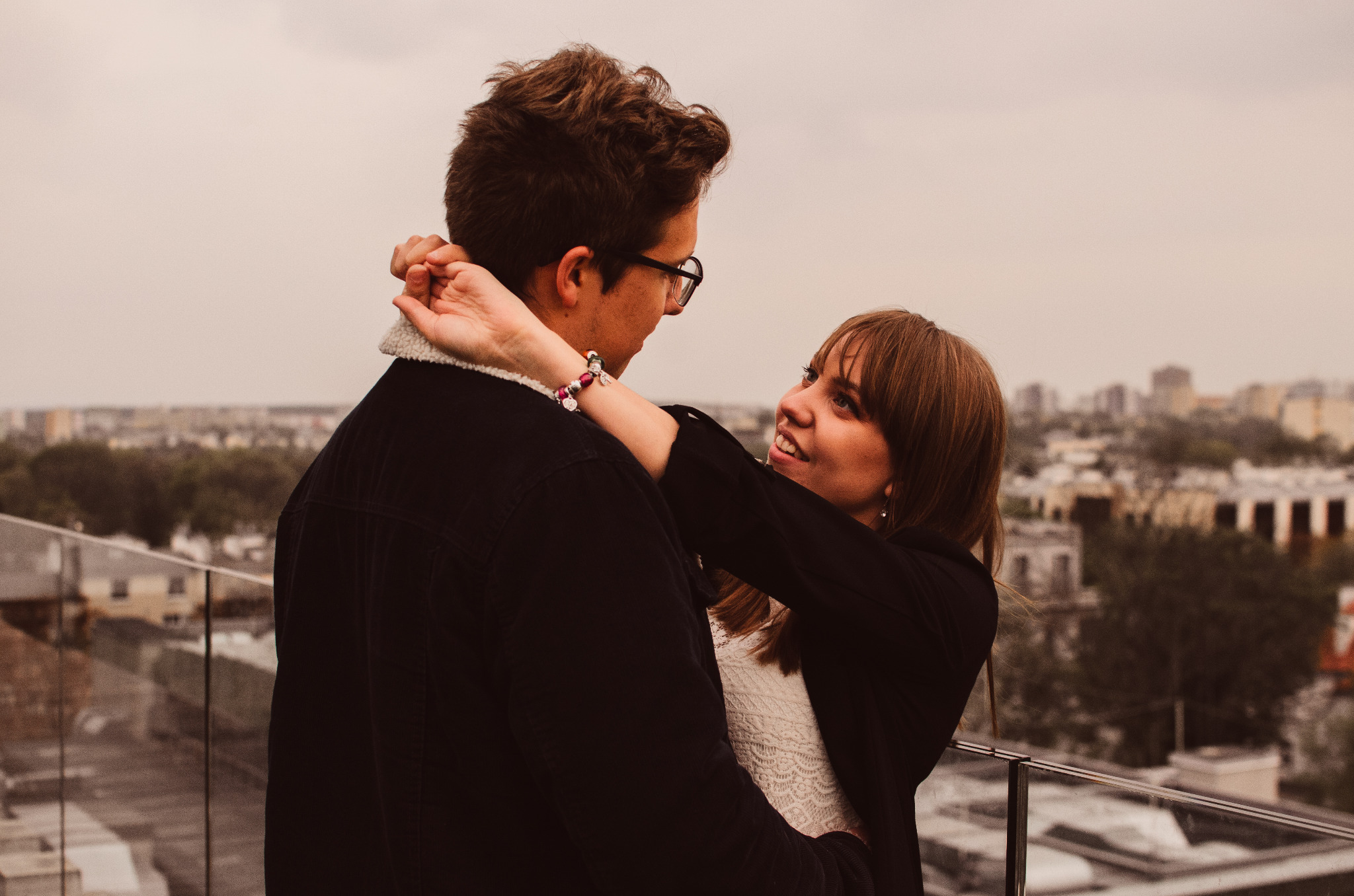 Para zakochanych, on w okularach i kurtce z barankiem, obejmowana przez uśmiechniętą kobietę na tle miejskiej panoramy z dachu budynku.