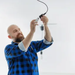 Brodaty mężczyzna w niebieskiej koszuli w kratę wymienia żarówkę LED w czarnej oprawie wiszącej na tle białego sufitu.