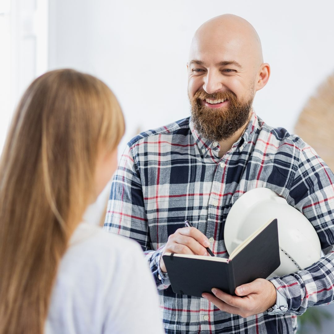 Uśmiechnięty mężczyzna z brodą w koszuli w kratę, trzymający kask ochronny i notatnik, rozmawia z klientką, prawdopodobnie omawiając zakres prac.