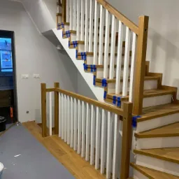 Montaż schodów: drewniane stopnie i białe balustrady zabezpieczone niebieską taśmą malarską, widoczne ślady prac wykończeniowych na podłodze.