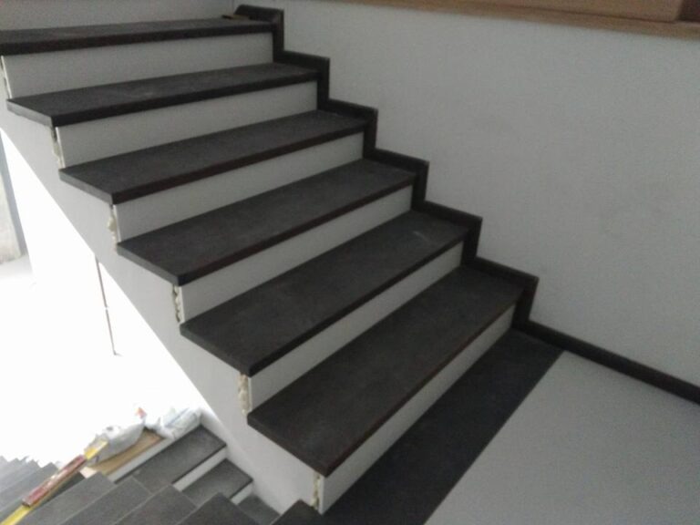 Montaż stopni z ciemnego drewna na białych schodach betonowych, widoczne pianka montażowa i płytki na podłodze.