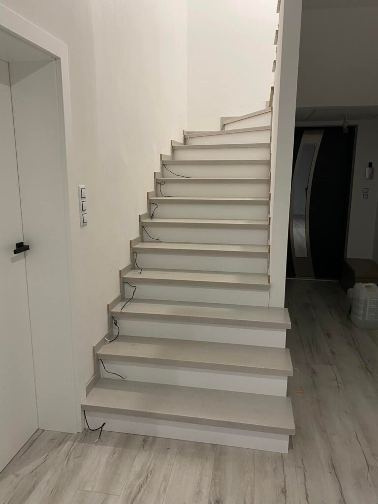 Wnętrze domu w trakcie wykańczania: jasne schody z podświetleniem LED w trakcie montażu, widoczne wystające kable, minimalistyczny design.