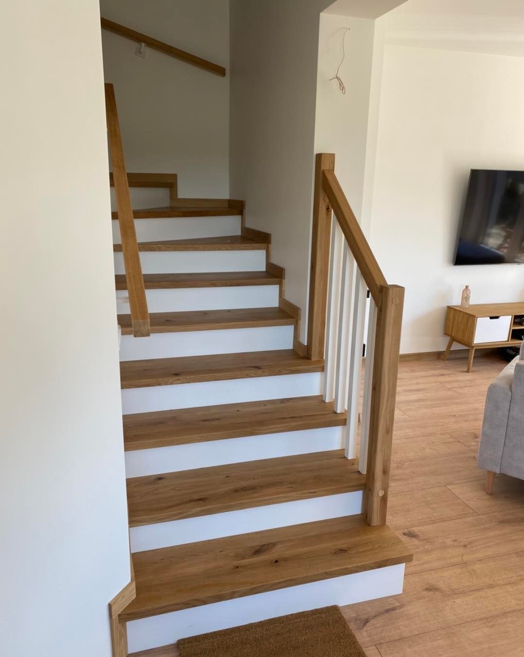 Drewniane schody z białymi podstopnicami i jasnym drewnem na stopniach, widok od dołu, nowoczesny design, jasne wnętrze z drewnianą podłogą i telewizorem w tle.