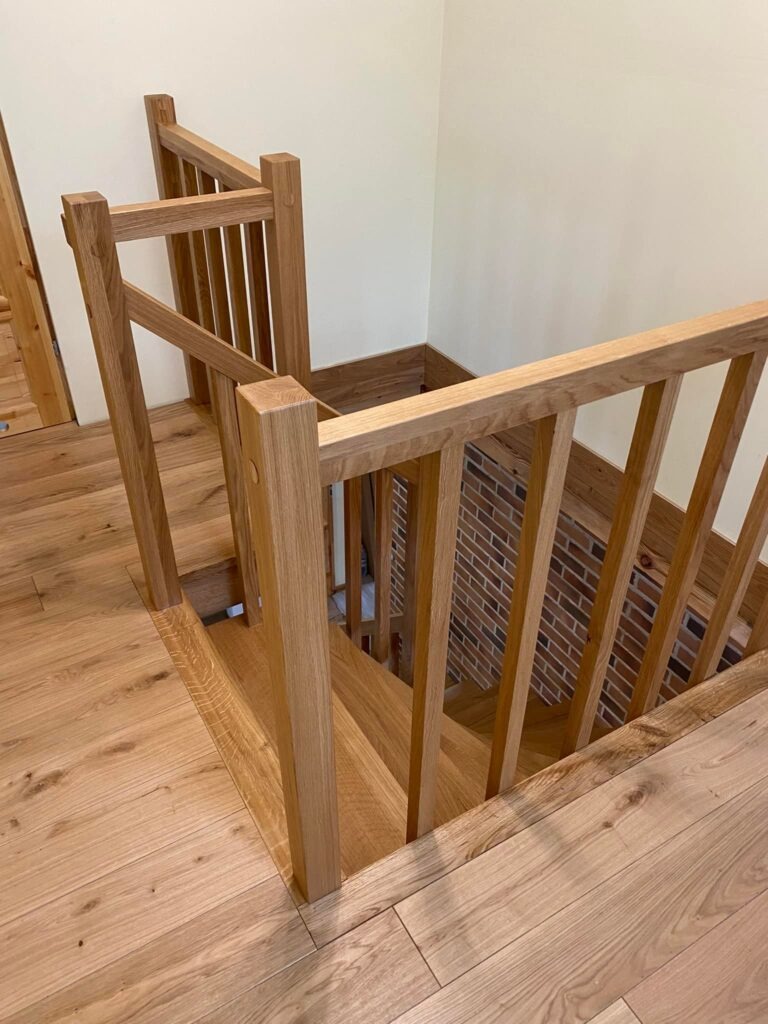 Jasne, dębowe schody z prostą balustradą, dopasowane do podłogi, prowadzące do piwnicy wykończonej cegłą.