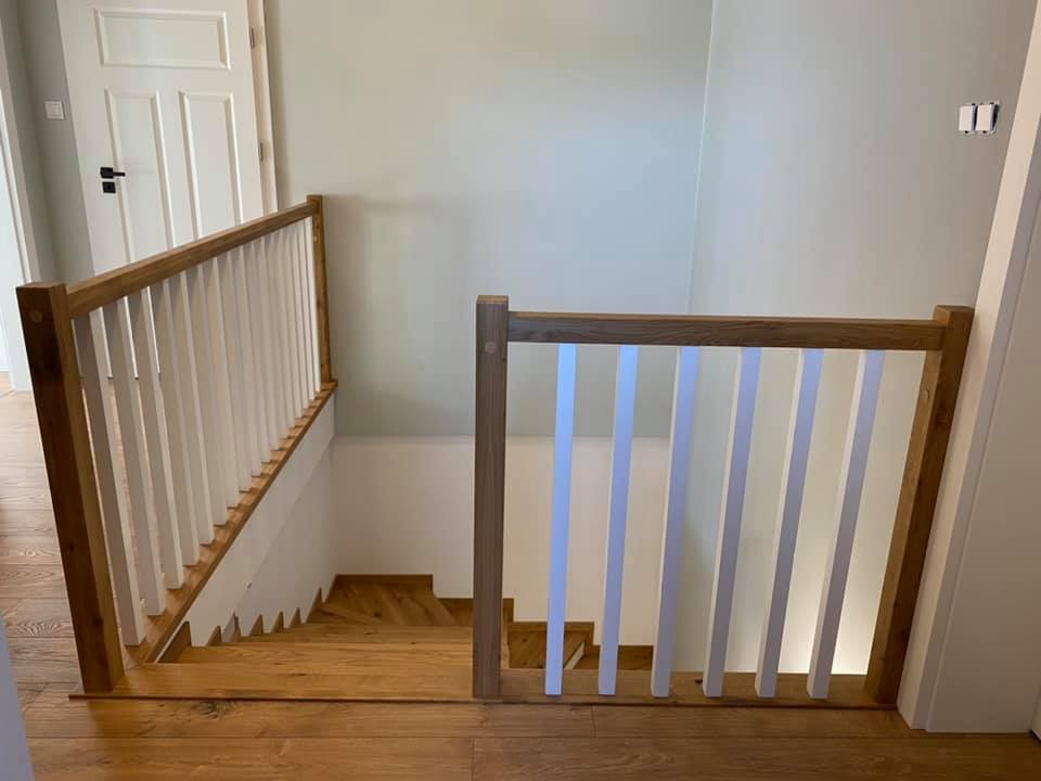 Jasne drewniane schody z białymi balustradami, widok z góry na dół, z widocznym fragmentem podłogi na górnym poziomie i białymi ścianami w tle.