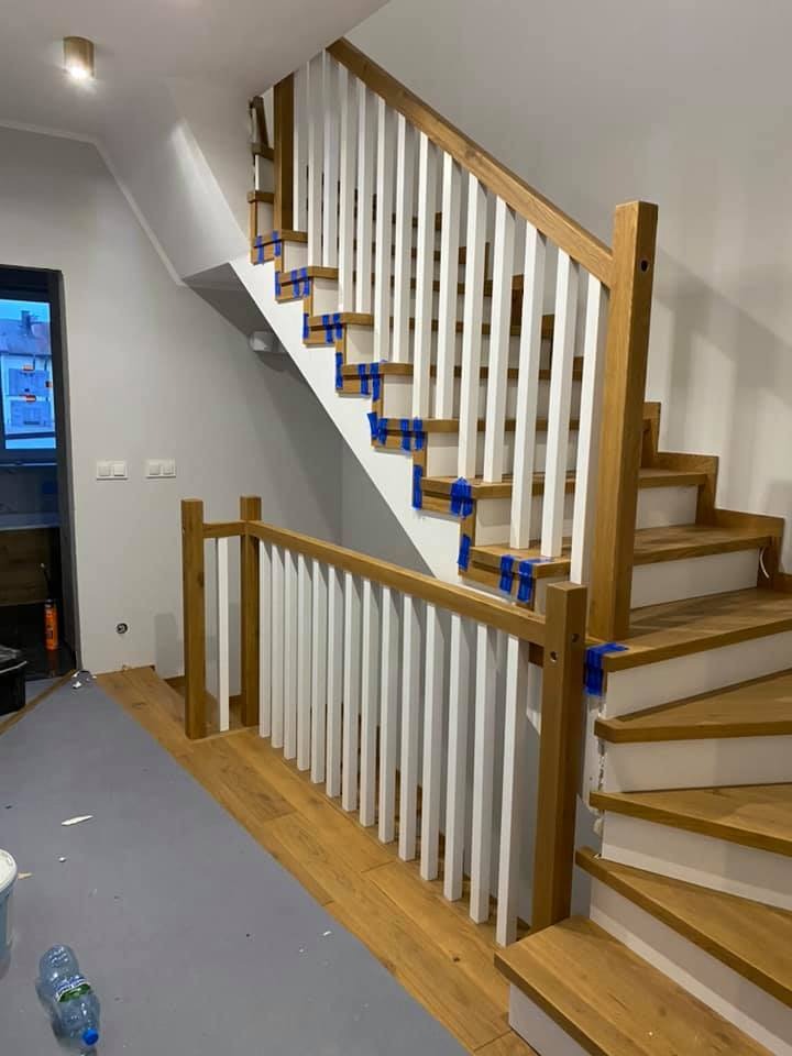 Montaż schodów: drewniane stopnie i białe balustrady zabezpieczone niebieską taśmą malarską, widoczne ślady prac wykończeniowych na podłodze.
