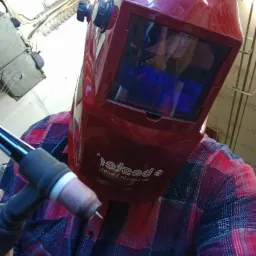 Osoba w czerwonej masce spawalniczej z automatycznym przyciemnianiem trzyma palnik TIG, ubrana w koszulę w kratę, przygotowuje się do spawania.