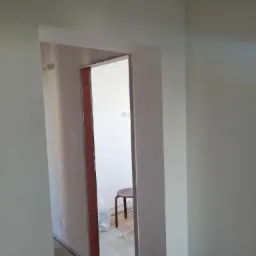 Świeżo pomalowane na szaro ściany w korytarzu, widok przez ościeżnicę do pokoju z krzesłem i workiem na śmieci, podłoga częściowo zabezpieczona folią.