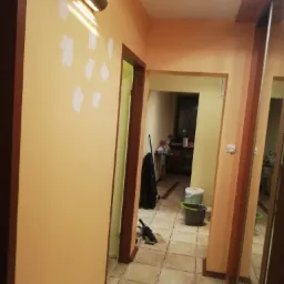 Korytarz w trakcie przygotowania do malowania, z widocznymi łatami gładzi na ścianie, podświetlonymi przez kinkiet, oraz sprzętem malarskim w tle.