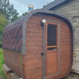 Drewniana sauna w kształcie beczki z brązowym gontem na dachu, częściowo ukończona, z widocznymi kablami i narzędziami wokół, umieszczona na betonowych blokach przed budynkiem.