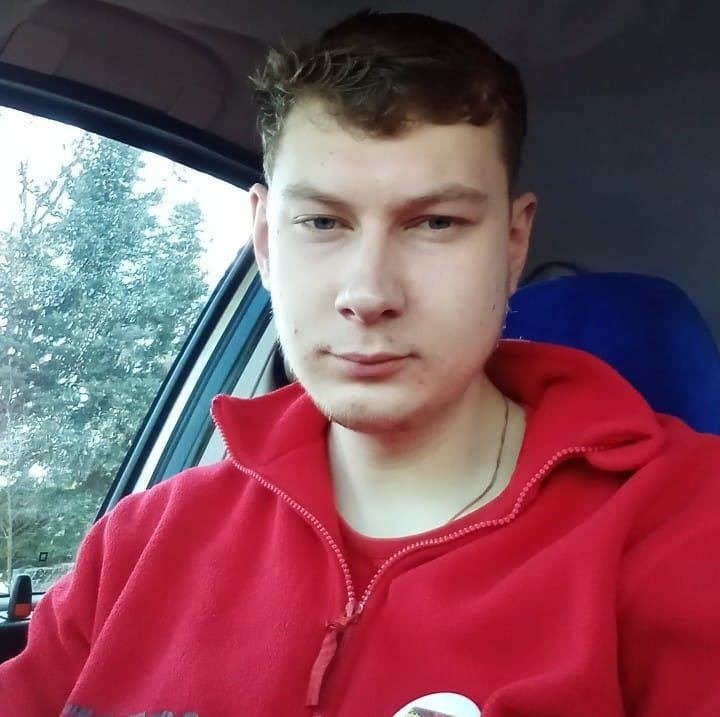 Portret mężczyzny w czerwonej bluzie z zamkiem błyskawicznym, siedzącego w samochodzie; widok przez okno na drzewa bez liści.