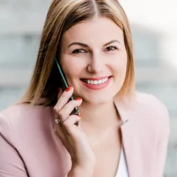 Uśmiechnięta blondynka w różowym żakiecie rozmawia przez telefon, prezentując manicure w kolorze czerwieni.