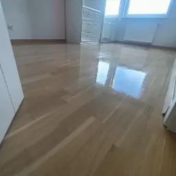 Błyszcząca, świeżo polakierowana podłoga z parkietu drewnianego w pokoju z białymi ścianami, widoczne odbicie okna w mokrej powierzchni lakieru.