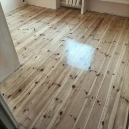 Jasna, drewniana podłoga po renowacji z widocznym, świeżo nałożonym lakierem, odbijającym światło. W tle widoczny grzejnik i wnęka w ścianie.