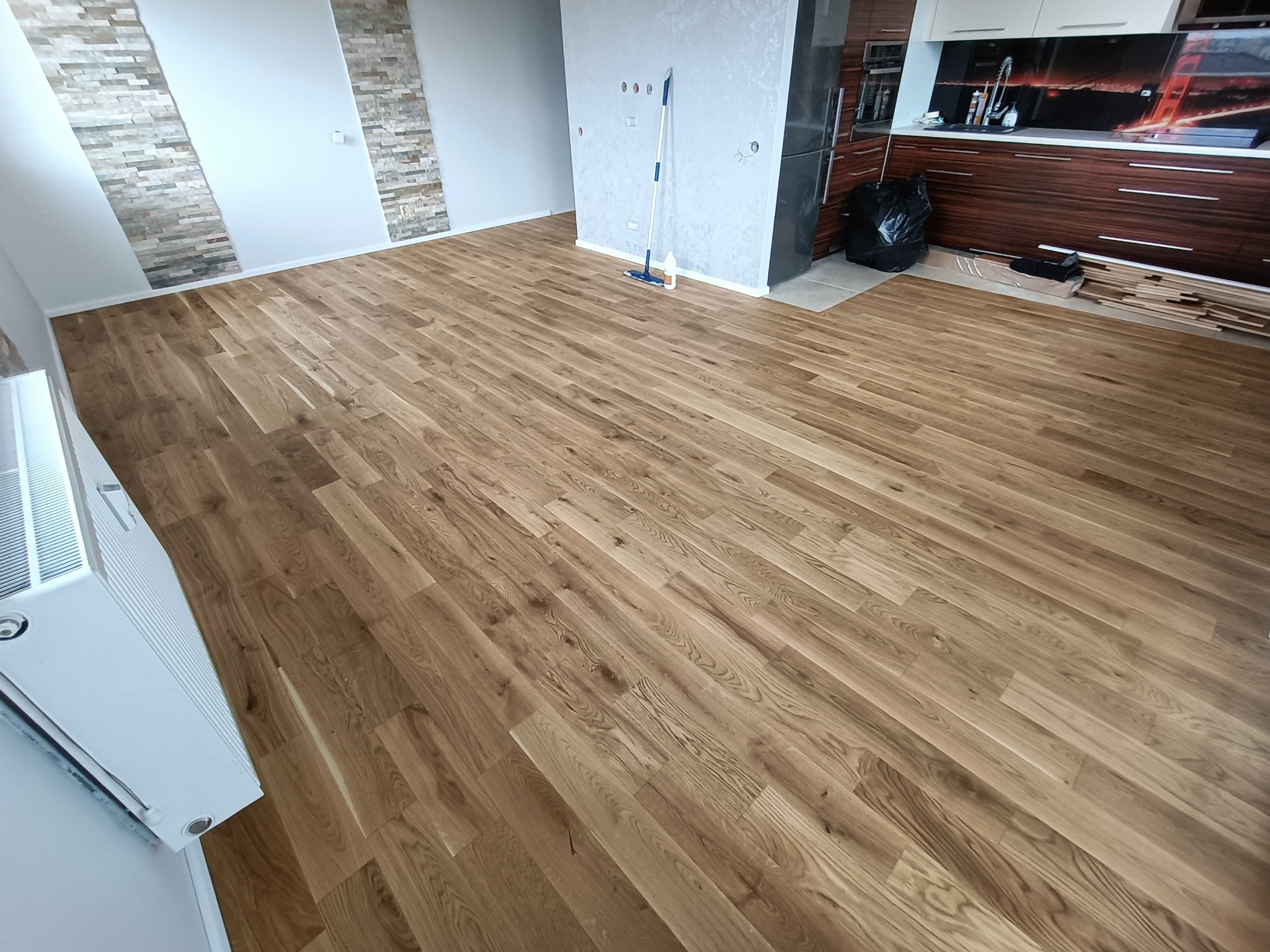 Jasny, drewniany parkiet ułożony w przestronnym salonie z dekoracyjną ścianą z kamienia, widoczny mop i butelka płynu do czyszczenia, częściowo widoczna zabudowa kuchenna z ciemnego drewna.