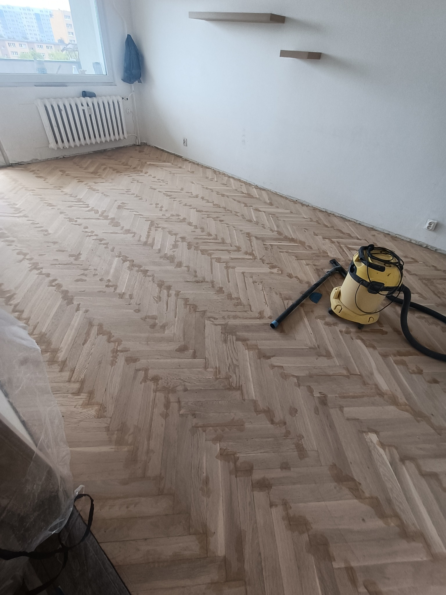 Parkiet ułożony w jodełkę, w trakcie czyszczenia, widoczny odkurzacz przemysłowy, ślady po czyszczeniu na drewnianej podłodze, w tle grzejnik i puste półki na ścianie.