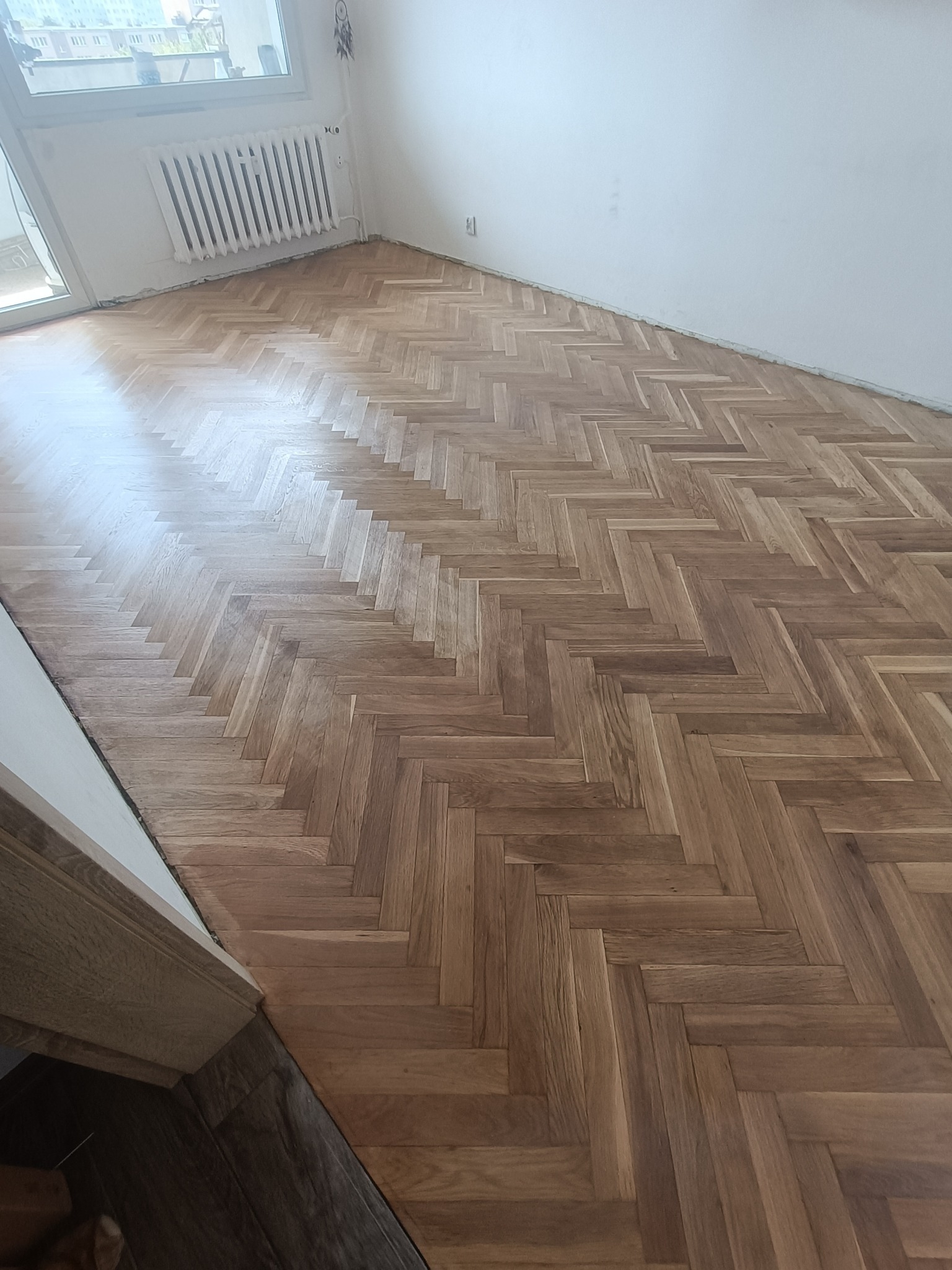 Ułożony parkiet w jodełkę w jasnym pomieszczeniu z białymi ścianami i grzejnikiem, widok z góry.