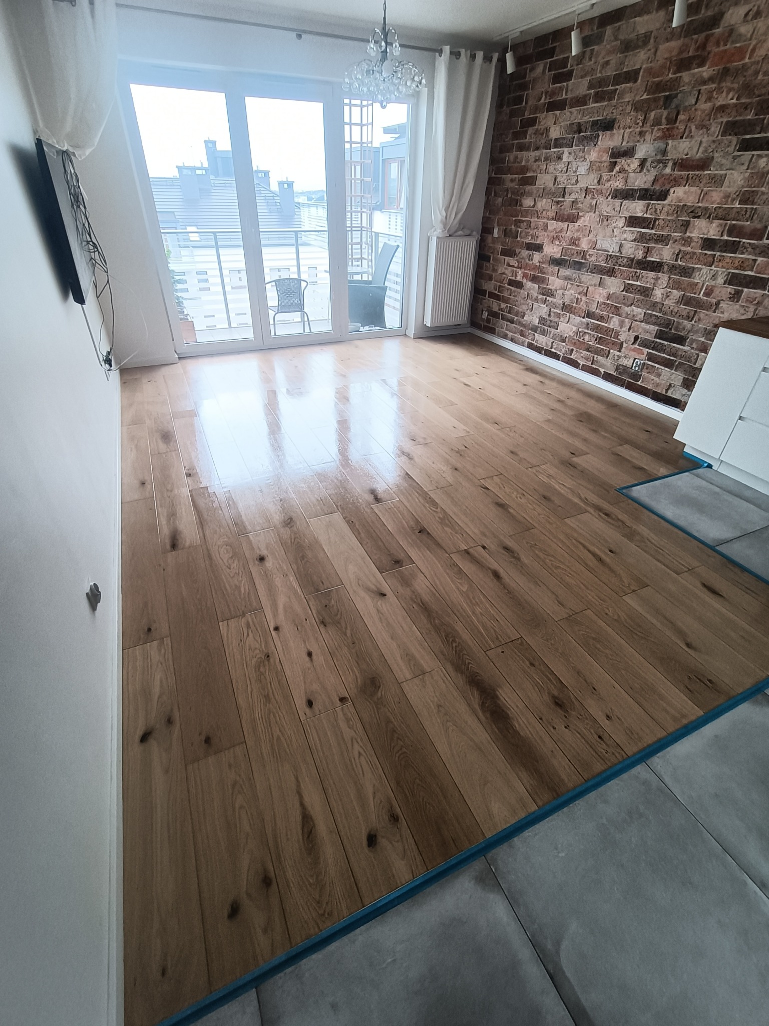 Jasny, drewniany parkiet ułożony w nowym mieszkaniu, widok na balkon i ceglaną ścianę, nowoczesne wykończenie.