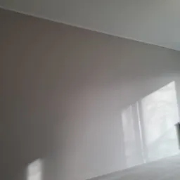 Świeżo pomalowane wnętrze w odcieniach szarości, z grzejnikiem i promieniami słońca wpadającymi przez okno, podkreślającymi gładką powierzchnię ściany.