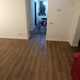 Ułożona, brązowa podłoga z paneli w nowo wyremontowanym pokoju, widoczny fragment czerwonej sofy i narzędzia w przejściu do kolejnego pomieszczenia.